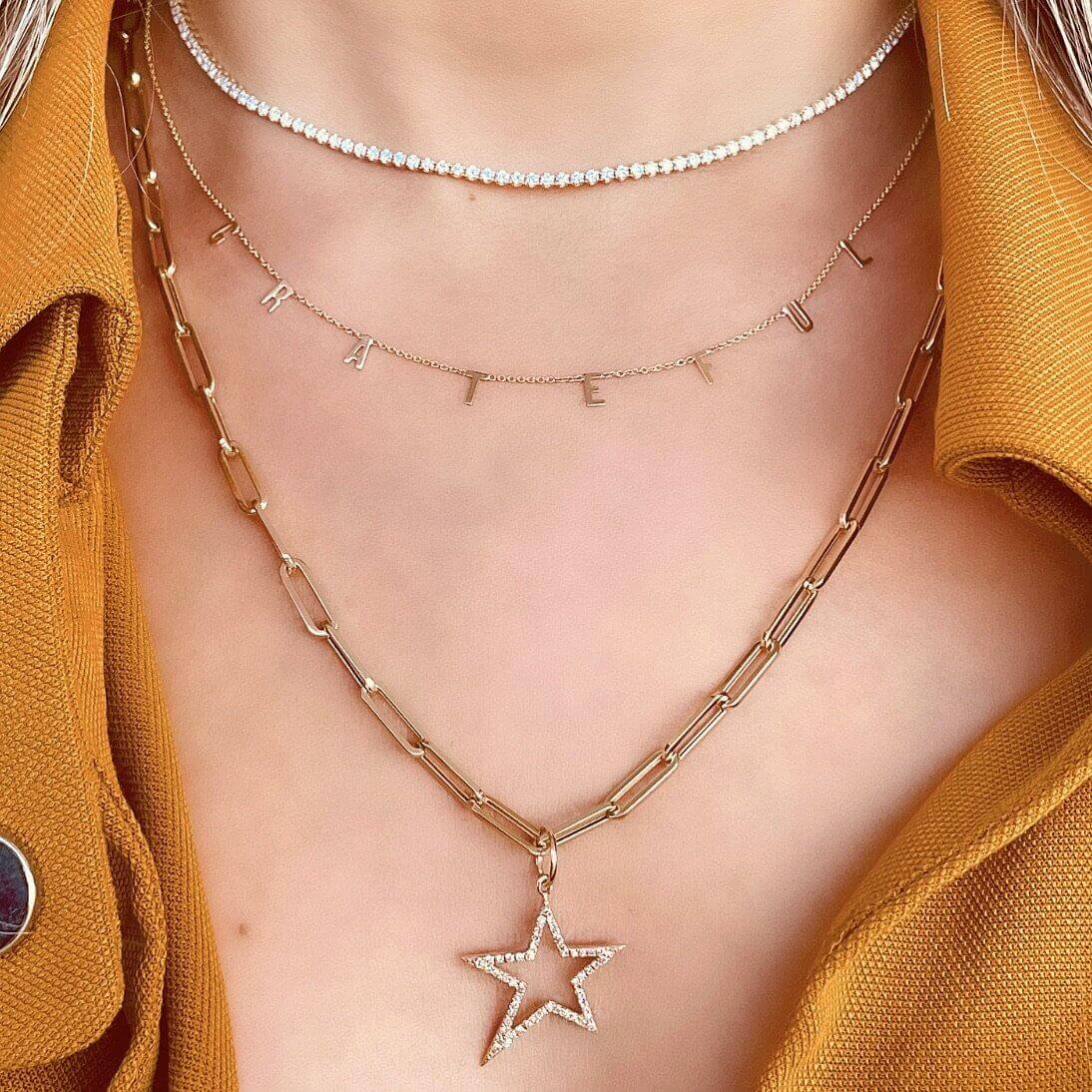 Layered necklaces featuring star pendant, letter charms, and beaded choker, styled with a mustard yellow top. Elegant and personalized jewelry.