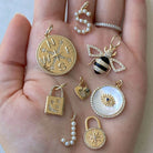 Hand displaying a collection of gold and gemstone pendants from our jewelry brand, featuring designs like a compass, bee, and evil eye.