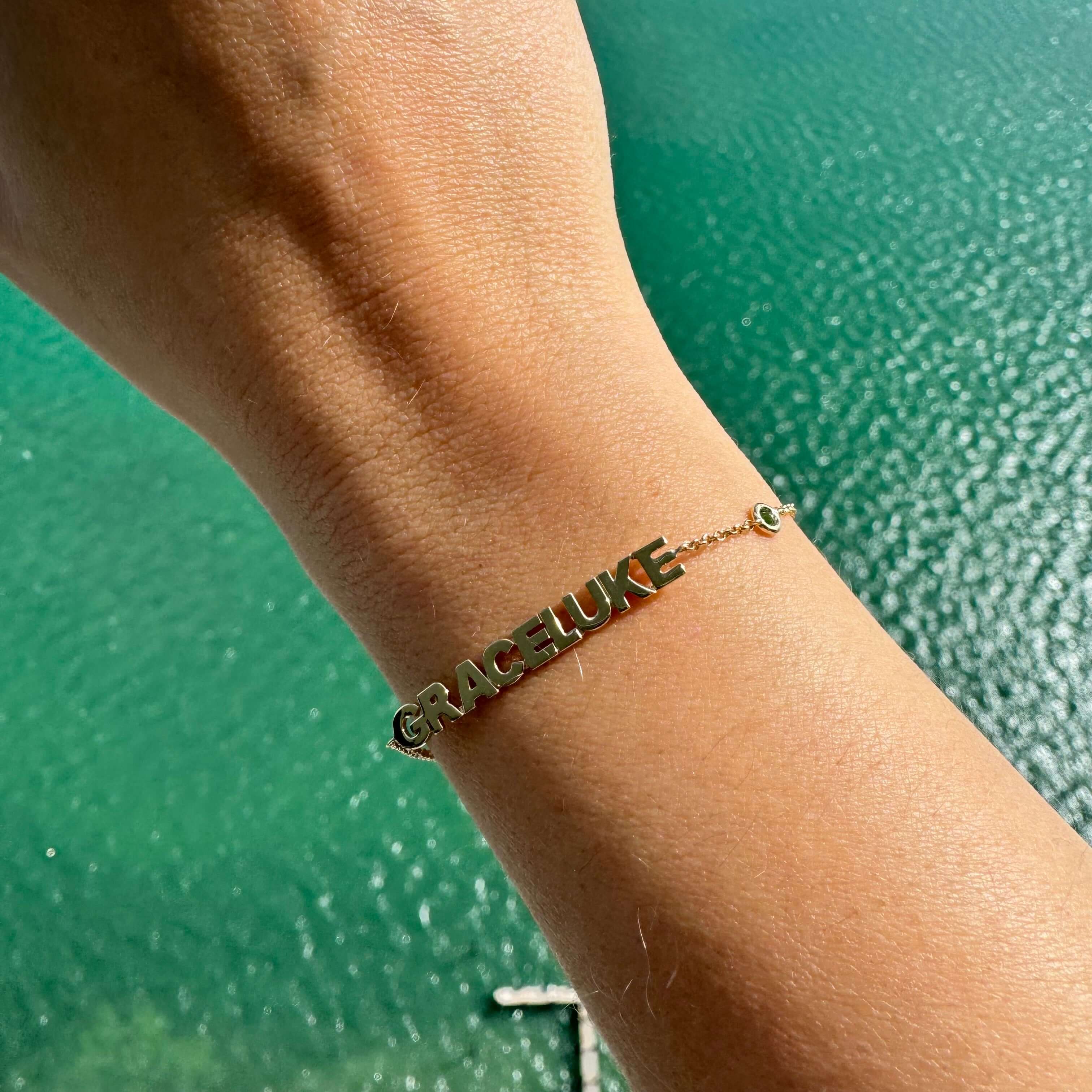 14K gold personalized block name bracelet featuring the names Grace and Luke against a turquoise water background.