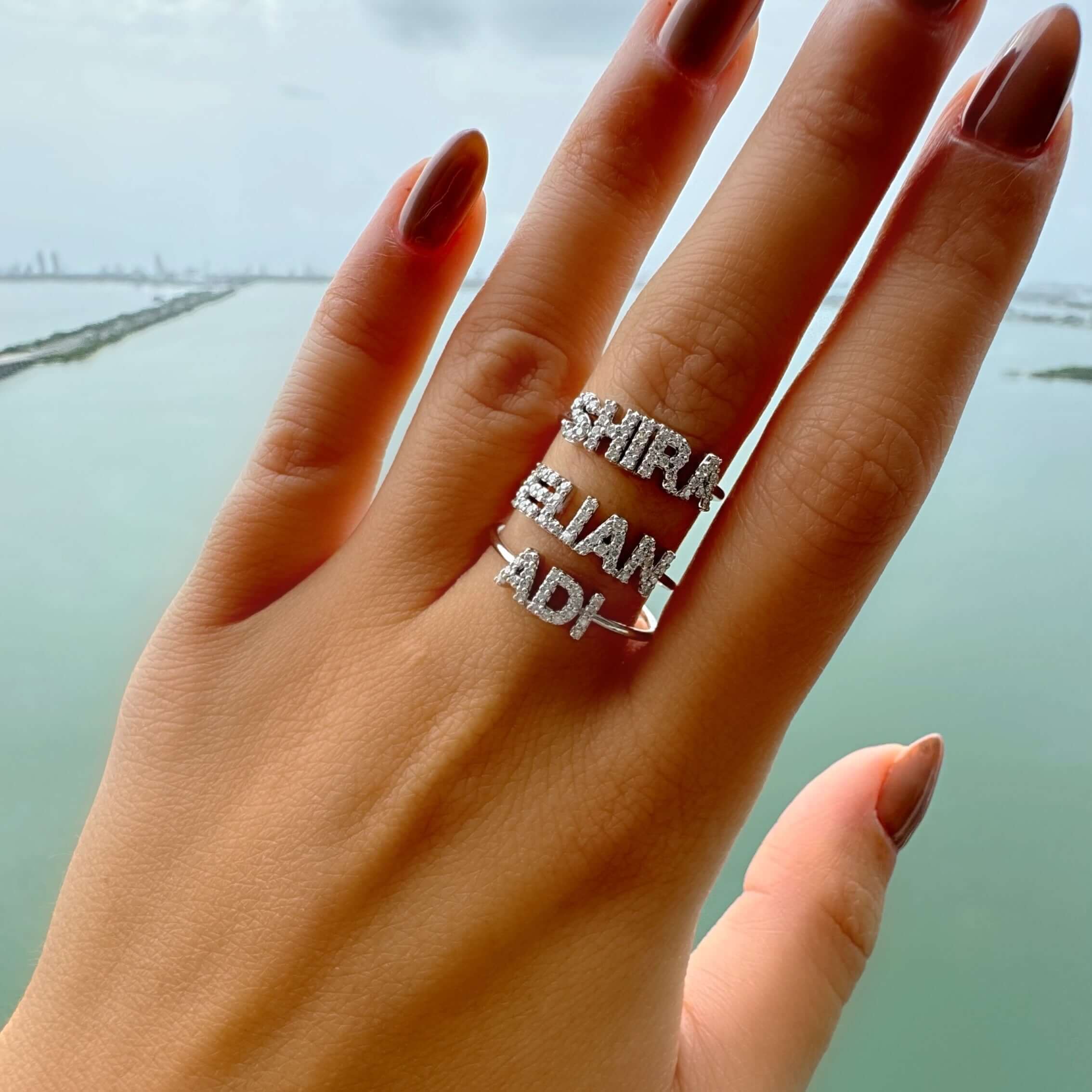 Woman's hand adorned with personalized silver name rings displaying coastal view, showcasing our custom jewelry collection.