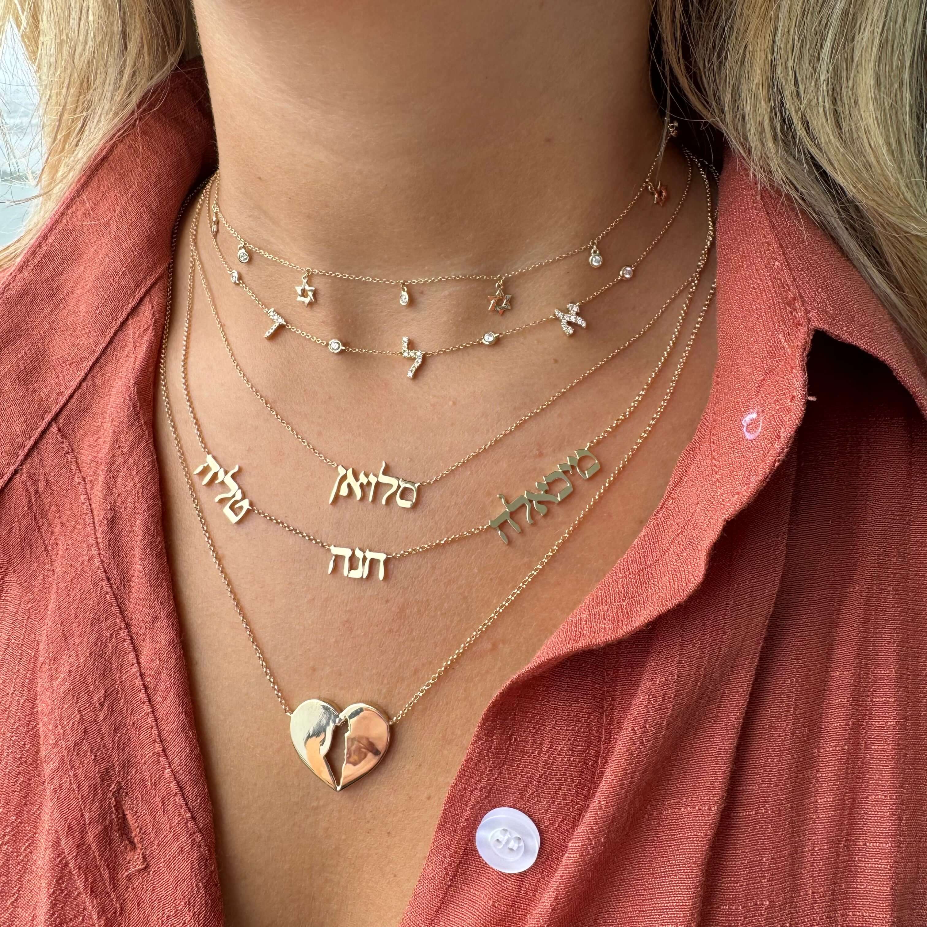 Woman wearing layered necklaces with Hebrew name pendants and heart charm, showcasing elegant jewelry design in a red blouse.