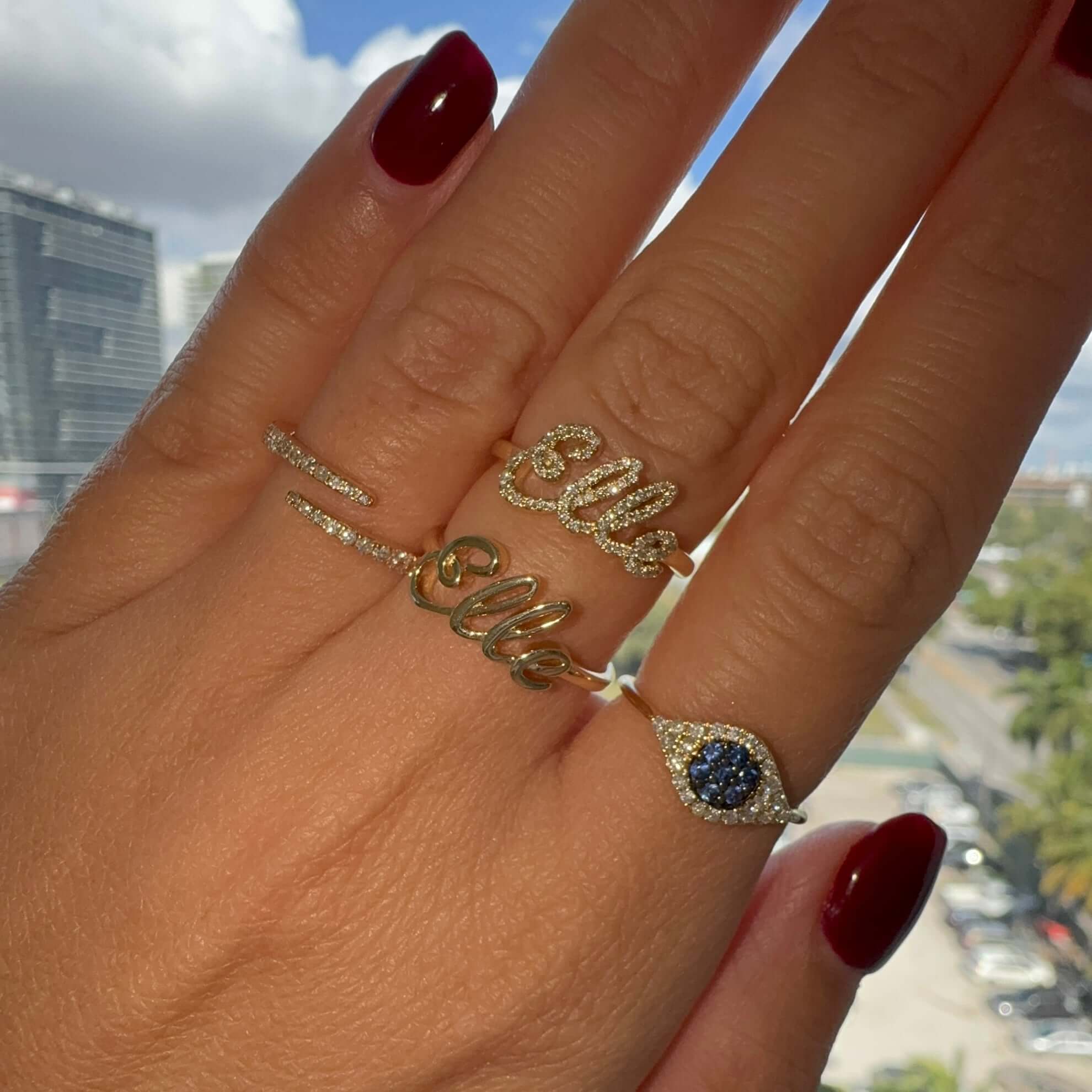 Close-up of a hand wearing multiple personalized gold diamond rings featuring the name "Elle" and a sapphire charm.