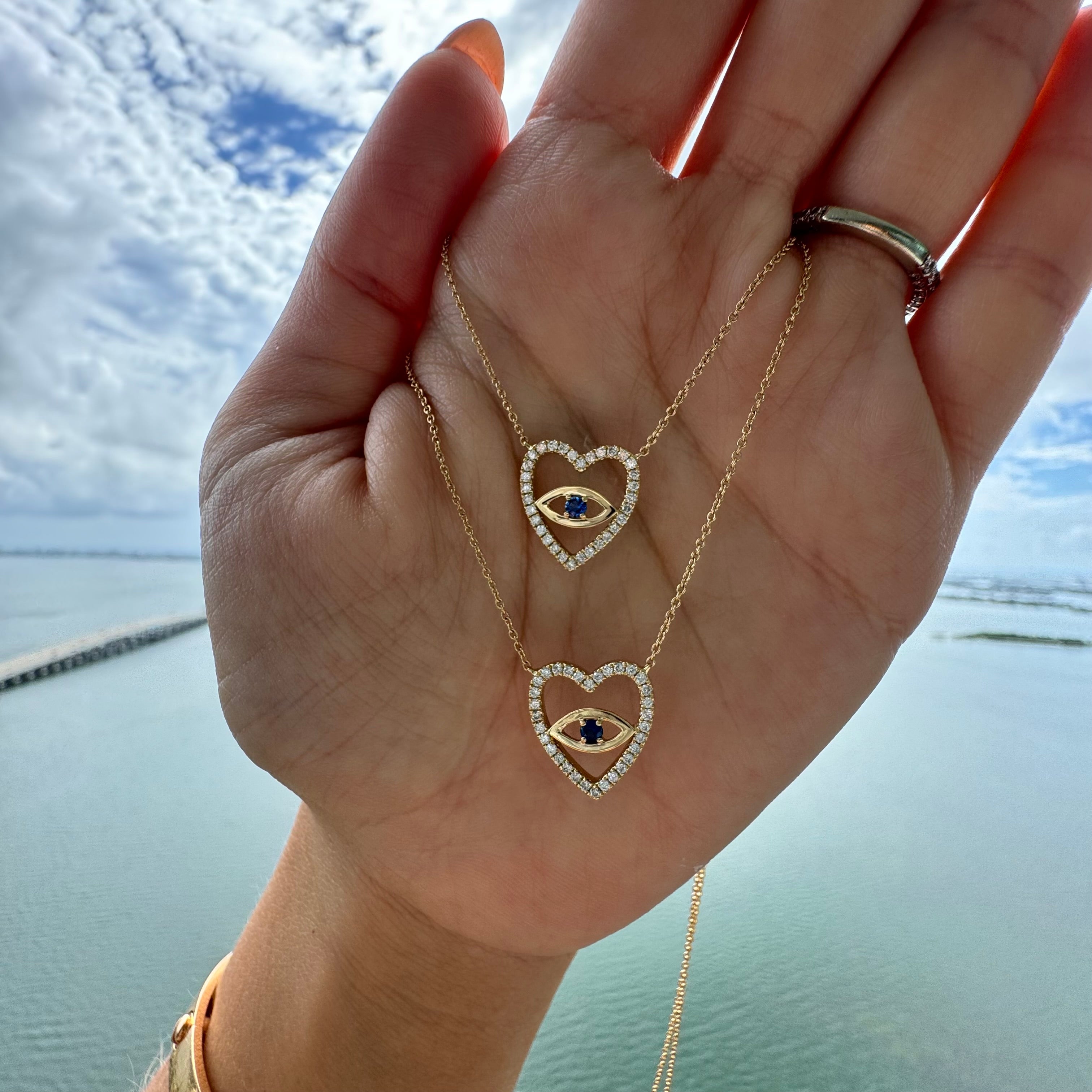 Hand holding two 14K gold heart necklaces with diamond accents, featuring sapphire evil eye designs, against a serene outdoor backdrop.