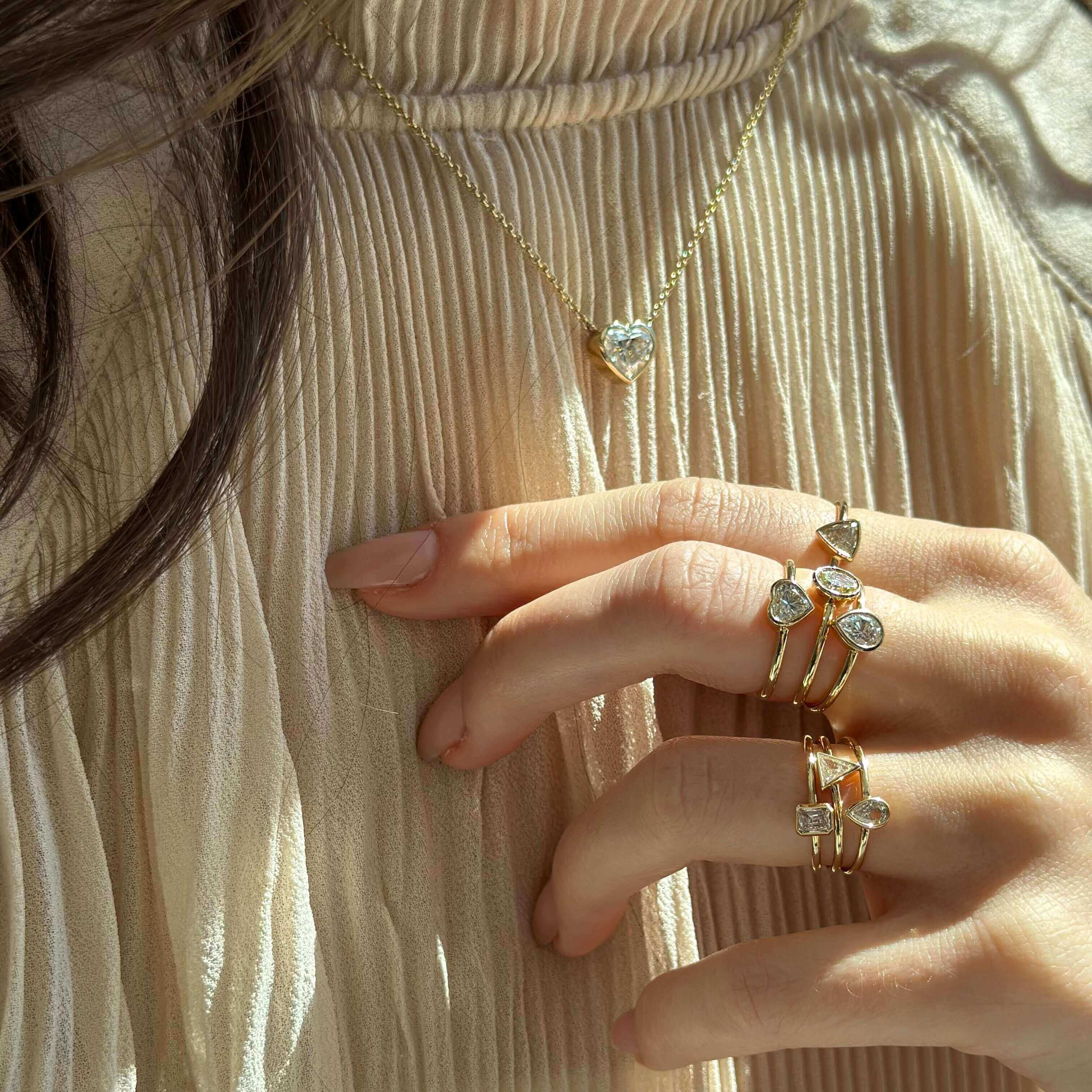 Woman's hand wearing gold rings and a heart necklace, showcasing elegant jewelry against a light textured fabric.