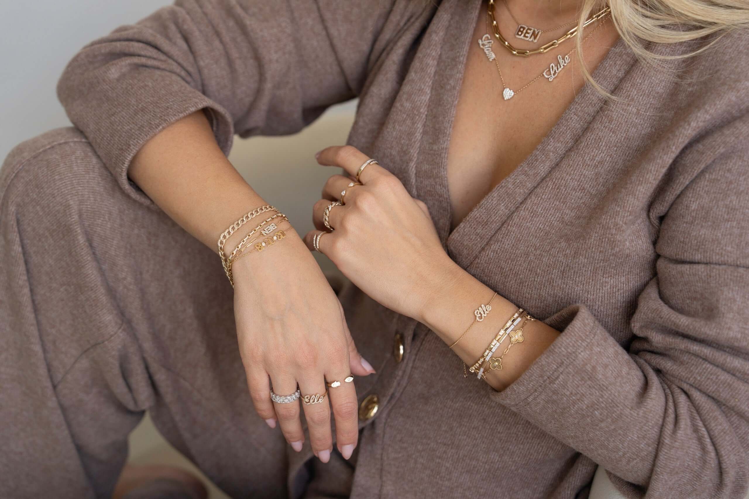 Woman showcasing elegant Izakov Fine Jewelry with layered bracelets and rings, dressed in a cozy outfit.