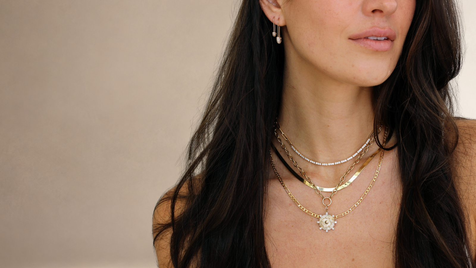 Woman wearing multiple gold necklaces and earrings against a neutral background