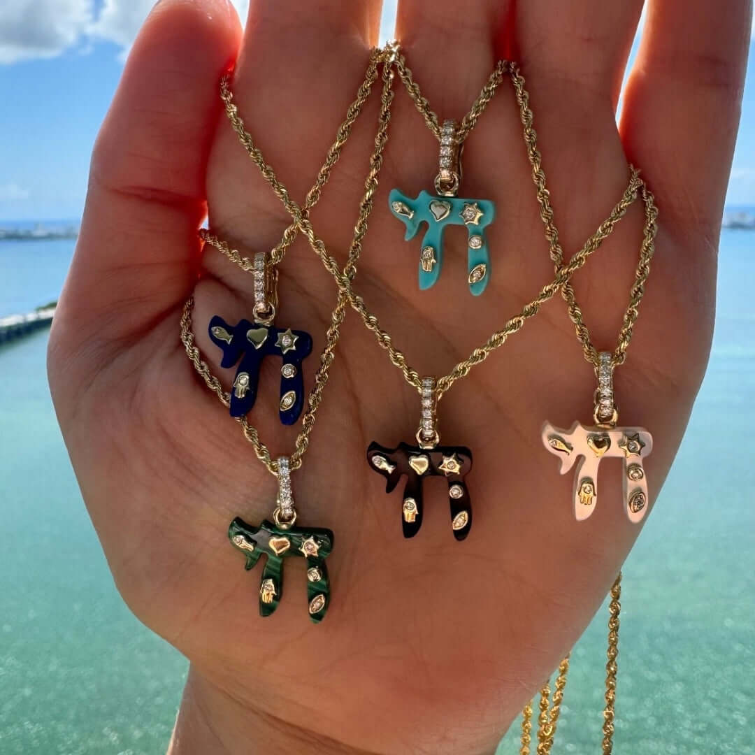 5 colorful 14K Gold Jewish Charms Chai Gemstone Pendant © displayed on a hand by the water, featuring various gemstones.