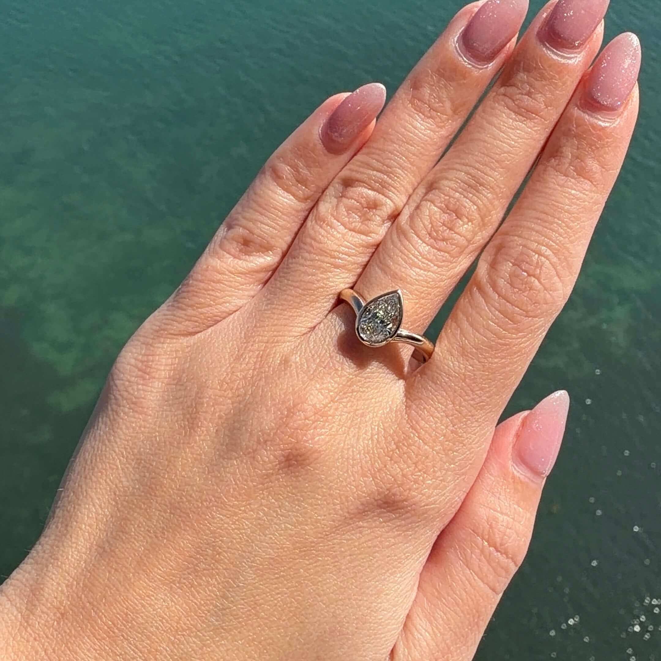 Curved Lab Diamond Bezel Ring worn on a hand with soft pink nails against a water background.